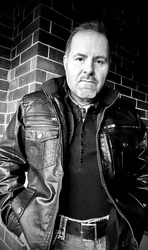 A man in leather jacket standing next to brick wall.